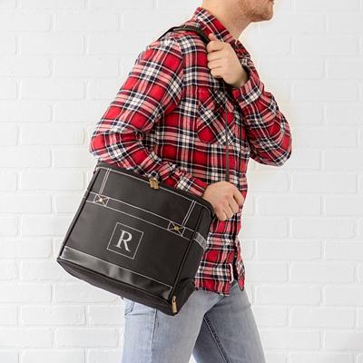 A person in a red plaid shirt and jeans holds the stylish black Back Door Brewski cooler tote with an R on it, set against a white brick wall, perfect for a groomsmen gift backdrop.