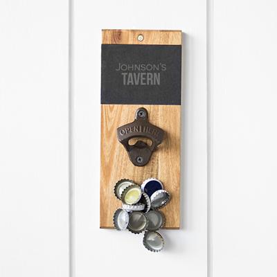 The wall-mounted Bar Blade, engraved with Johnsons Tavern, is perfect as a groomsmen gift. It features a cap catcher to collect bottle caps below and is crafted from wood and metal, hanging elegantly on a white wall.