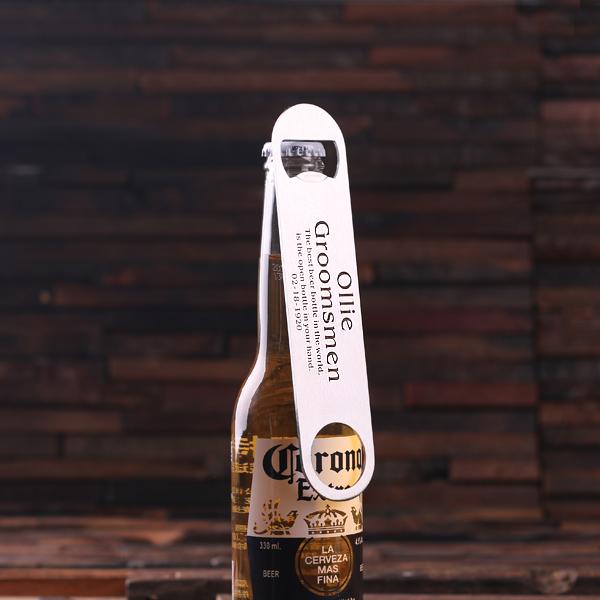 A Corona beer bottle with a stainless steel Aw Snap opener featuring personalized engraving reading Ollie Groomsmen and a date sits against a textured, dark brown wall. Ideal for any beer enthusiast.