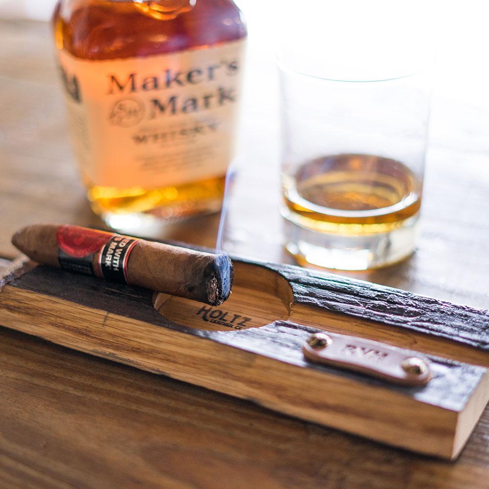 An Ash and Whiskey cigar holder, complete with a partially smoked cigar, rests on the table. Nearby, a glass of whiskey accompanies an open Makers Mark bottle. The rustic ashtray has a brass plaque and captures the charm of aged whiskey barrels.
